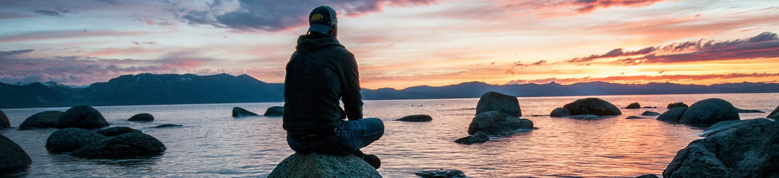 the psychology of decision-making, man sitting on rock staring out at water at sunset